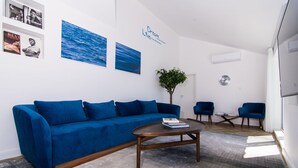 Un salon avec un canapé bleu, une table basse et deux fauteuils. On y trouve également des œuvres d’art murales, une horloge et une plante.