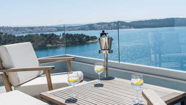 Un balcon offrant une vue dégagée sur un lac et sur le paysage urbain au loin. On y trouve deux chaises blanches avec des coussins et une table en bois sur laquelle reposent deux verres contenant une boisson au citron.