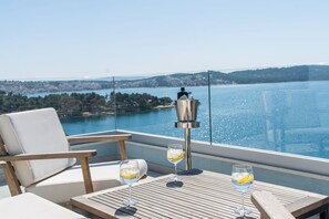 Un balcon offrant une vue dégagée sur un lac et sur le paysage urbain au loin. On y trouve deux chaises blanches avec des coussins et une table en bois sur laquelle reposent deux verres contenant une boisson au citron.