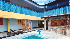 A swimming pool with a dolphin statue, a covered patio with blue lattice, and a stone wall with a wooden door. - Pousada Aquários do Porto (Ipojuca)
