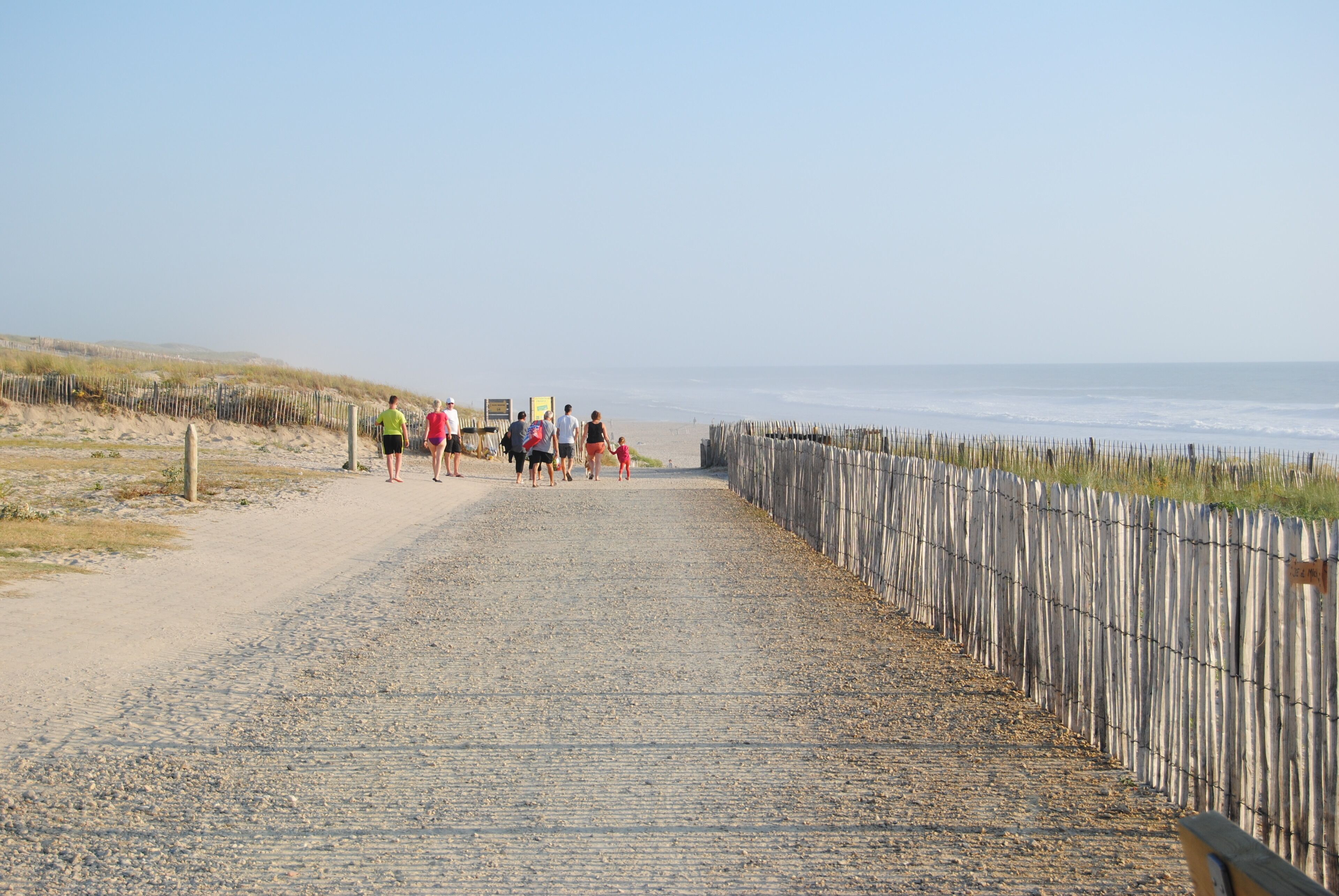 Ein gepflasterter Weg am Strand, eine Seite begrenzt von einem Holzzaun, die andere Seite von einer Grasdüne.