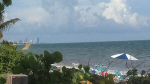 A beach with umbrellas and lounge chairs, a city skyline in the background, and a cloudy sky. - Paradise Found, Unique Location (Key Biscayne)
