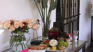 A table with a variety of food items, including cheese, fruits, and flowers, arranged on a glass table against a white wall with a metal gate in the background. - Plumbago Guest House (Mbombela)