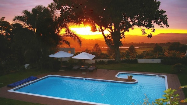 A rectangular swimming pool with a small hot tub, surrounded by a brick deck and a grassy area. There are two umbrellas and lounge chairs. - Plumbago Guest House (Mbombela)
