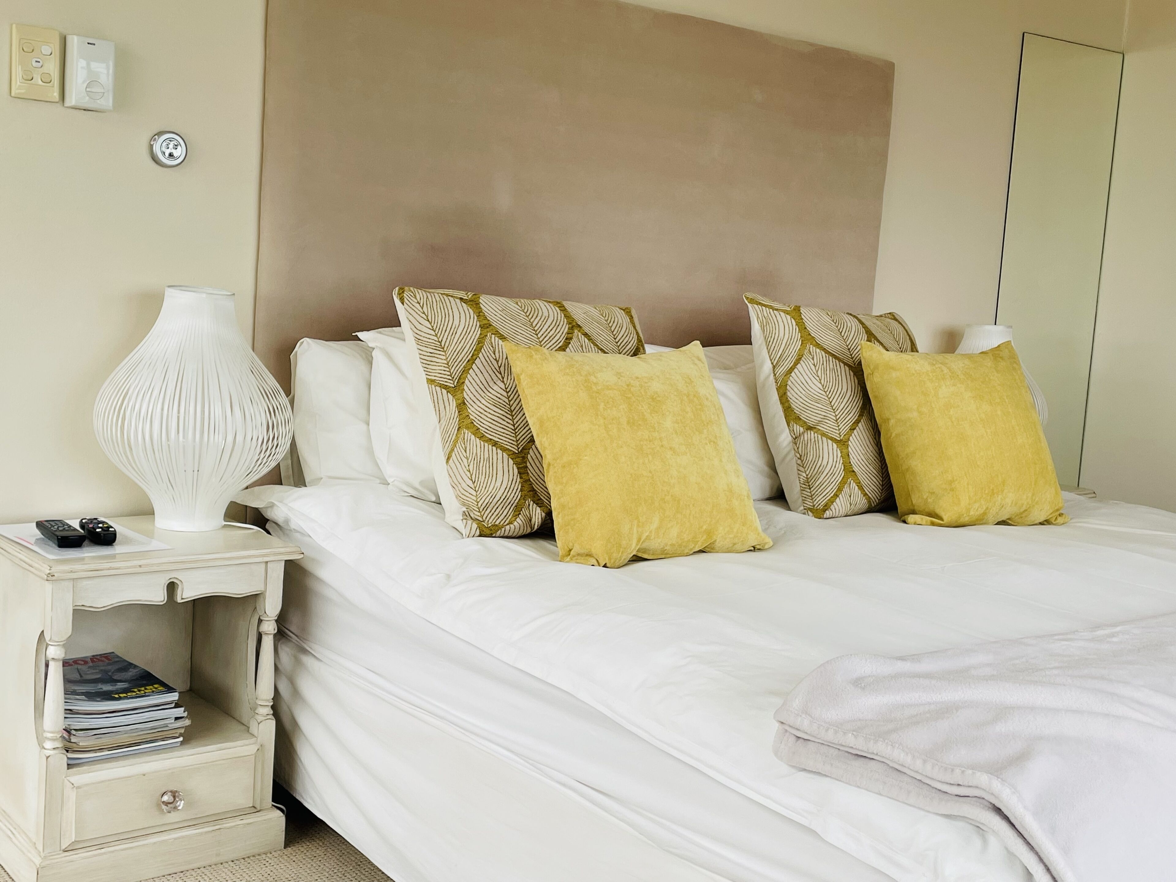 A neatly made bed with white bedding, a bedside table with a lamp, and a nightstand with magazines.