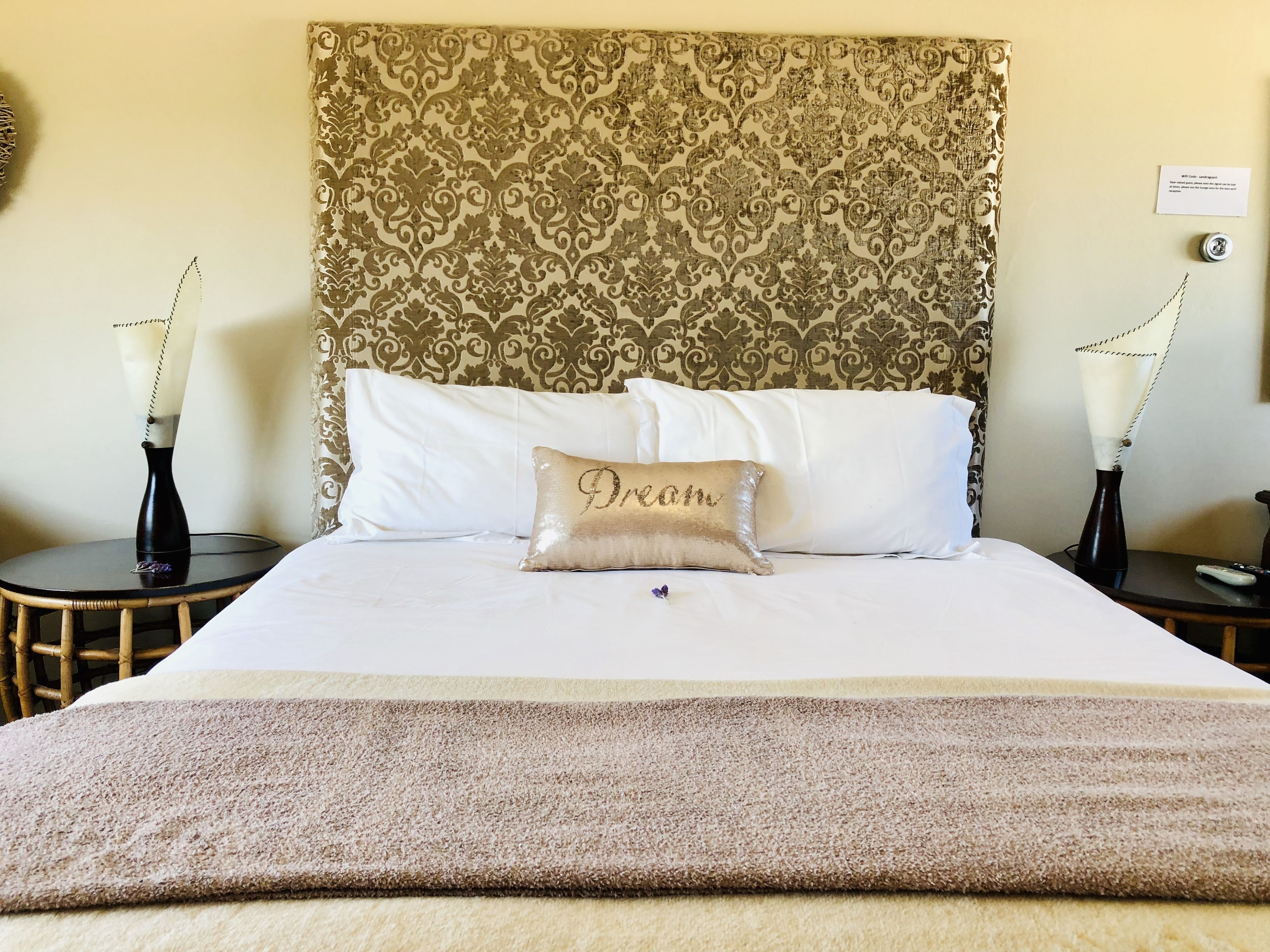 Uma cama bem arrumada com um travesseiro 'Dream', cabeceira estampada e duas mesas de cabeceira com vasos.