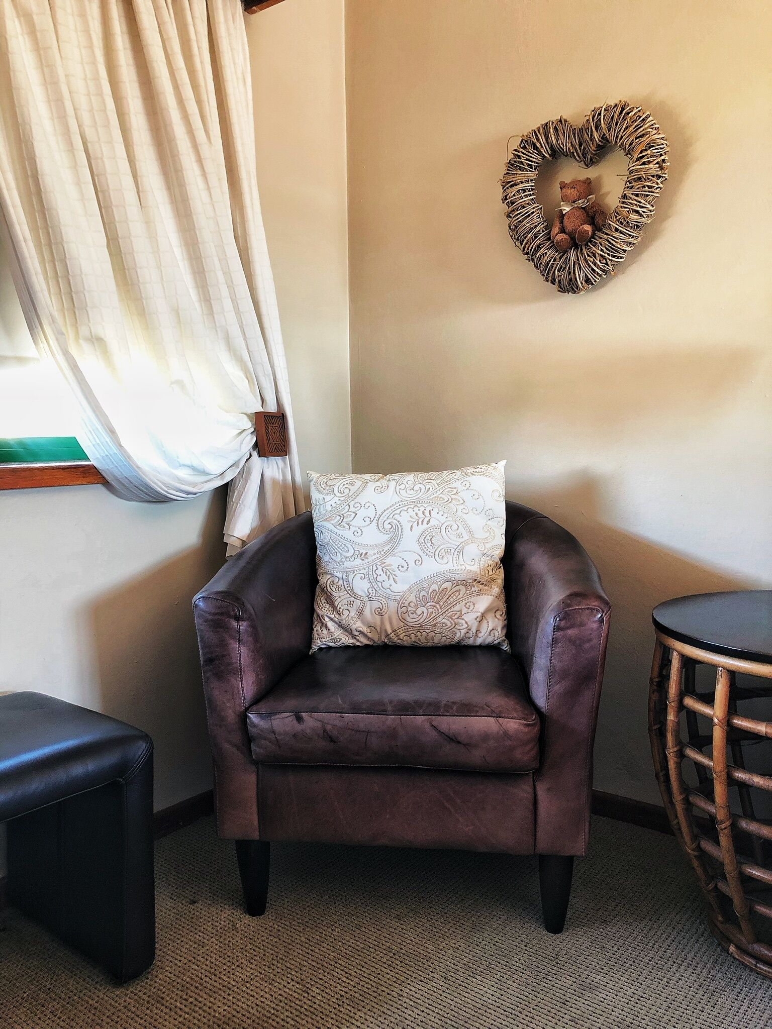 A corner room with a brown leather armchair, a patterned throw pillow, a heart-shaped wall decoration, a small round table, and a window with curtains.
