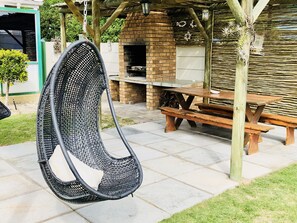 Ein Hängekorbstuhl, ein Backstein-Barbecue und ein Picknicktisch in einer Gartenanlage.