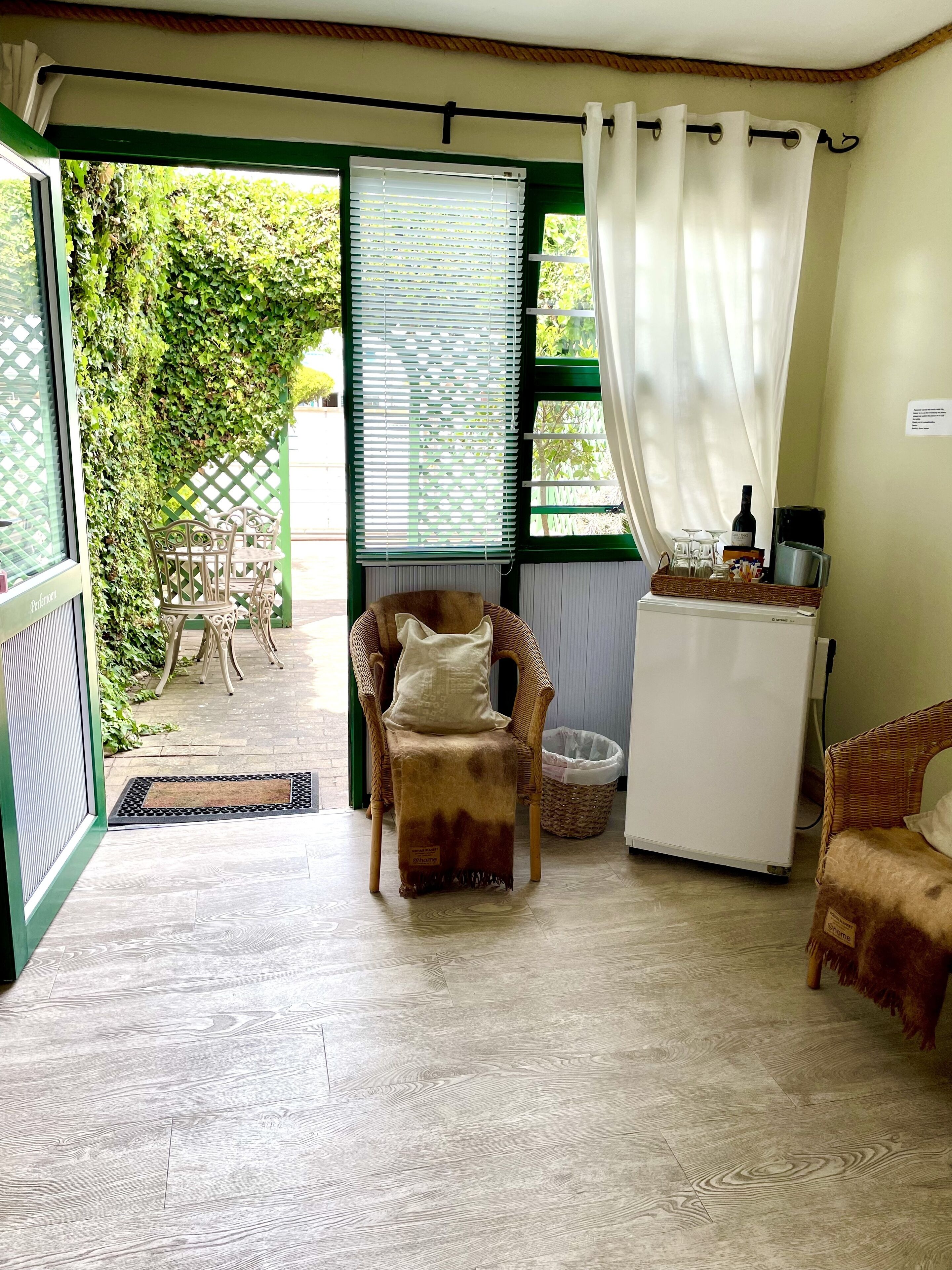 A cozy room with a green-framed window, a wicker chair, a small refrigerator, and a wicker sofa.