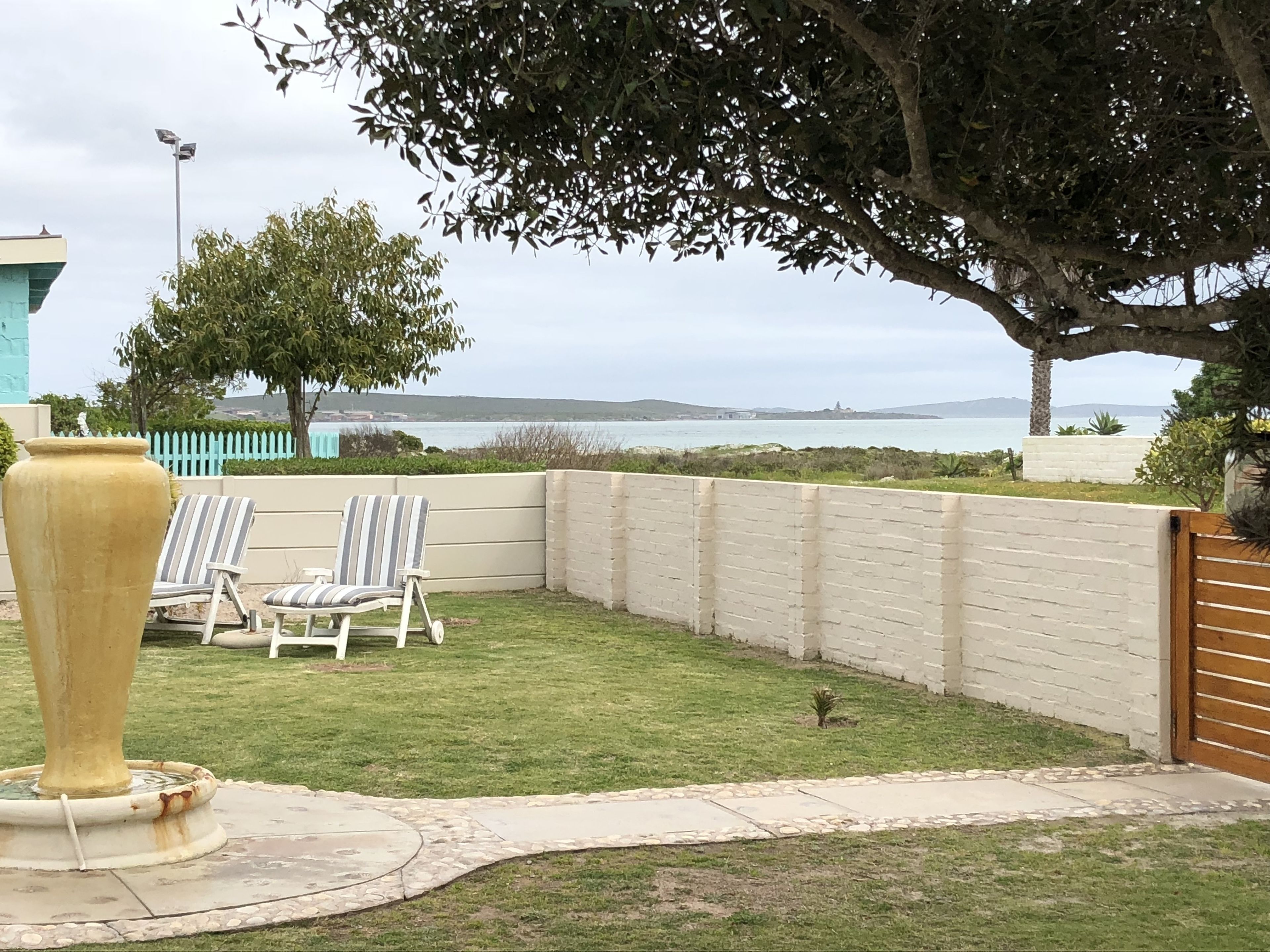 A backyard with a grassy lawn, two striped lounge chairs, a large yellow vase, and a view of the sea and hills in the distance.