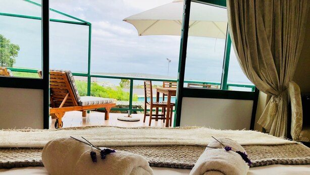 A hotel room with a large window, a view of the outdoors, a bed with two rolled towels, a wooden chair, and a small table with an umbrella. - Sandra's Guest House (Langebaan)