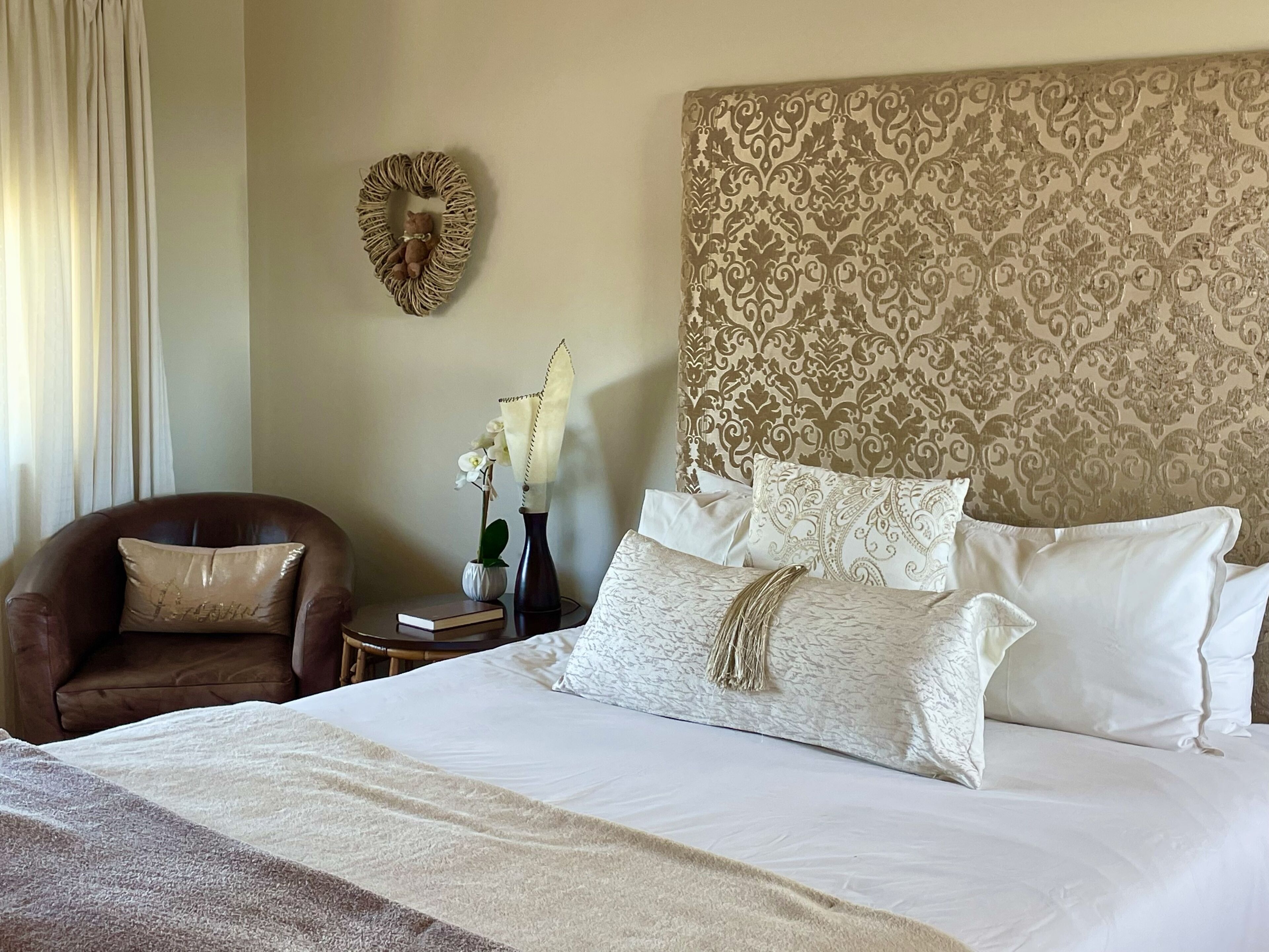 A neatly arranged bedroom with a large bed, a brown leather armchair, a small bedside table with a vase of flowers, and a decorative wall piece.