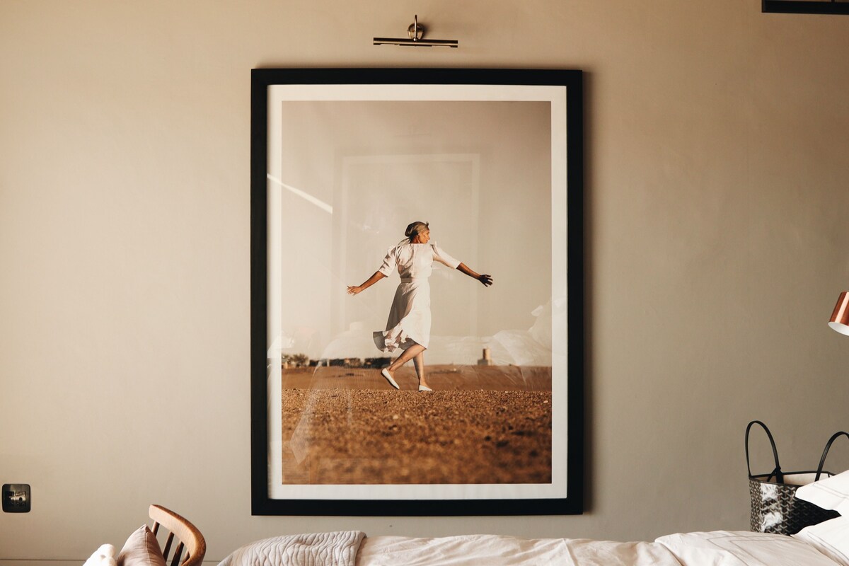 A framed photograph of a person in a field, a bed with white bedding, and a wooden chair with a cushion.