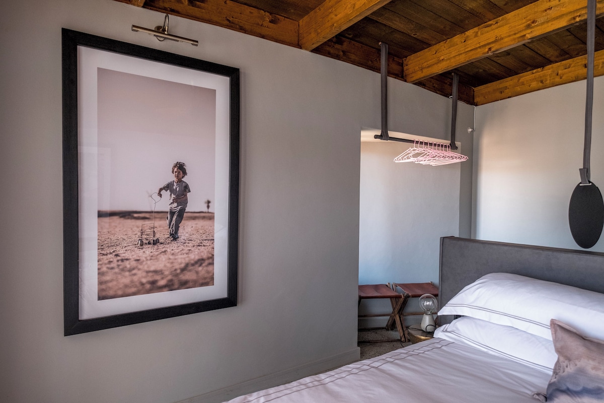 A bedroom with a bed, a framed photograph on the wall, a bedside table, and a hanging light fixture.