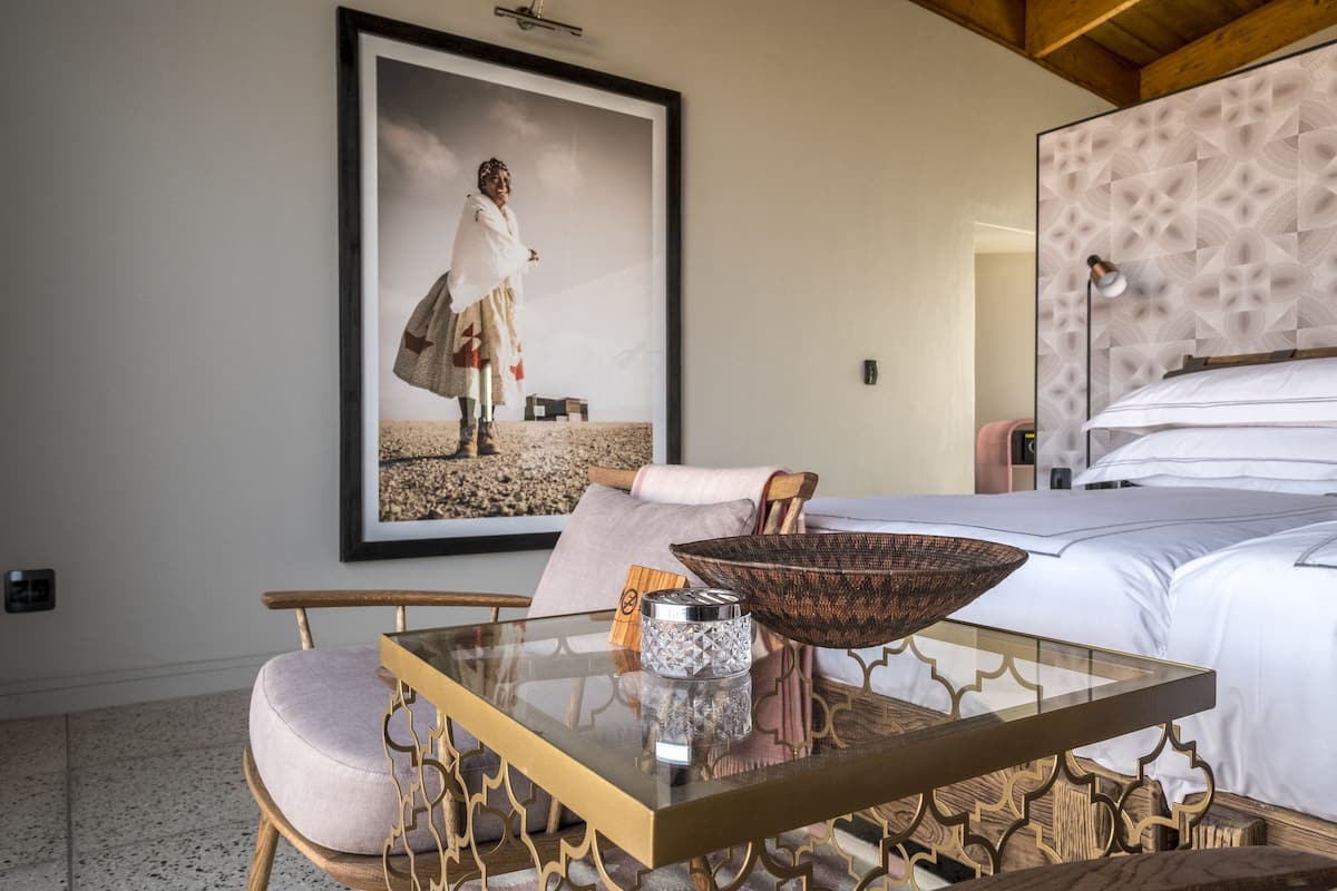 A hotel room with a bed, a glass-top table with a decorative bowl, a framed picture on the wall, and a wooden chair.