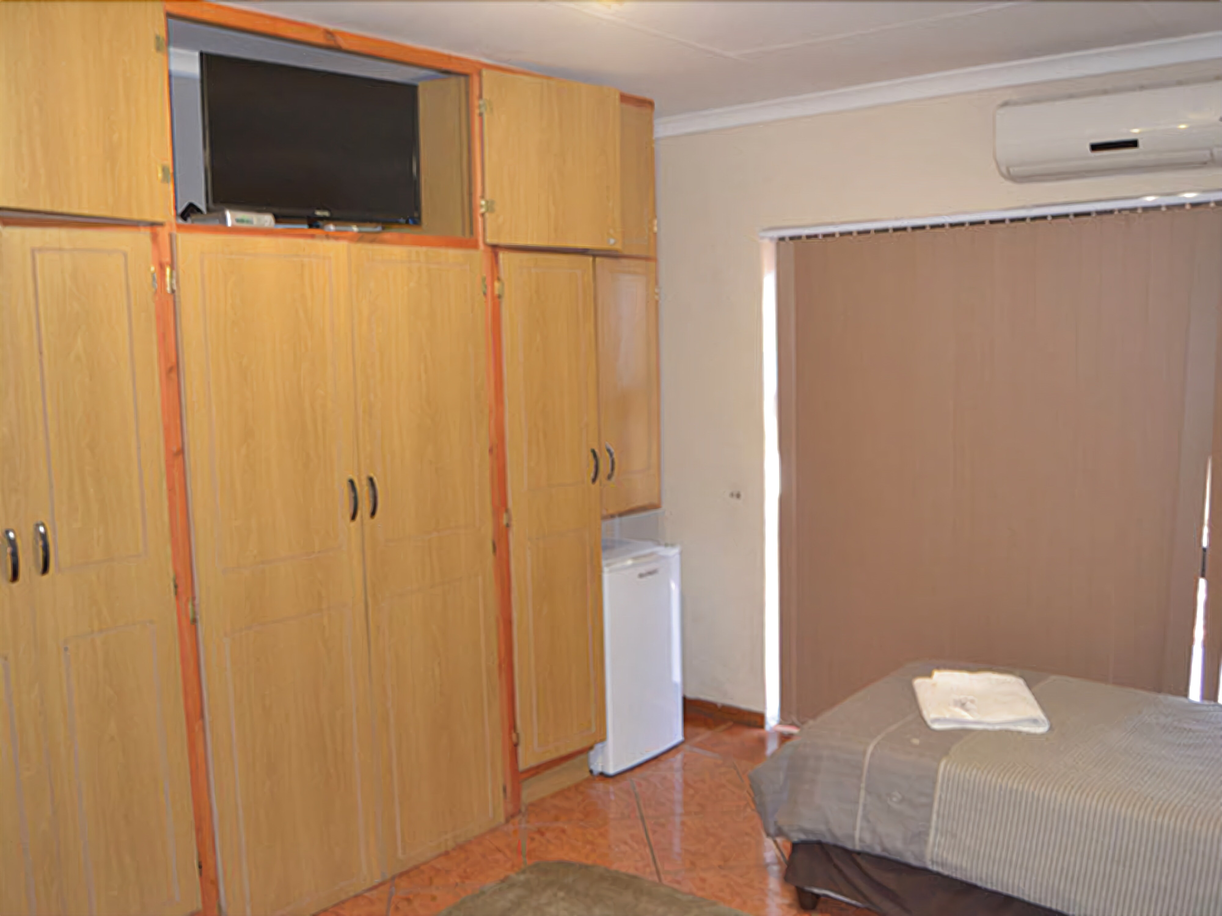 Une chambre avec des armoires en bois, une télévision, un petit réfrigérateur et un lit avec une couverture.