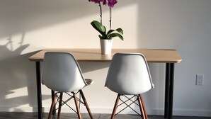Una habitación moderna con una mesa de madera y dos sillas blancas con patas de cobre. Hay una planta en maceta con flores moradas sobre la mesa.
