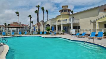 A pool area with blue lounge chairs, a sign reading "BEACH CLUB", and a building with a balcony and a tower-like structure.