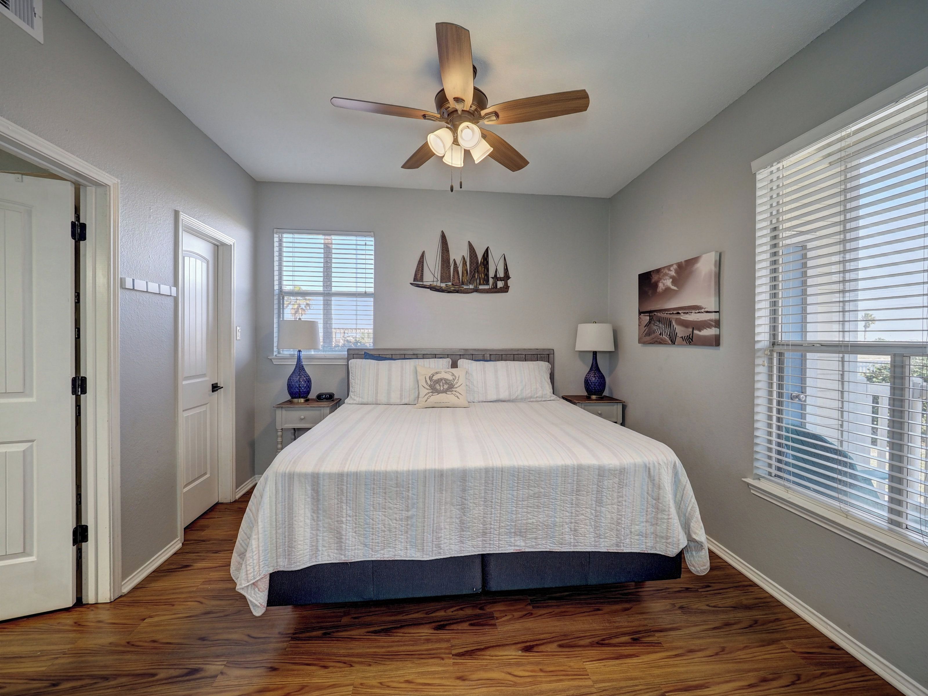 A bedroom with a bed, a ceiling fan, a nightstand with a lamp, a window with blinds, and a door.