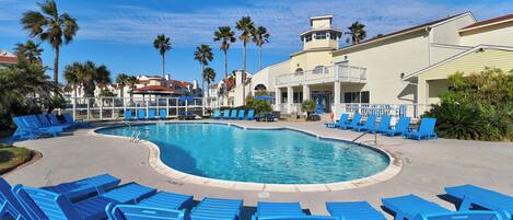A pool area with blue lounge chairs, palm trees, and a building with a tower.