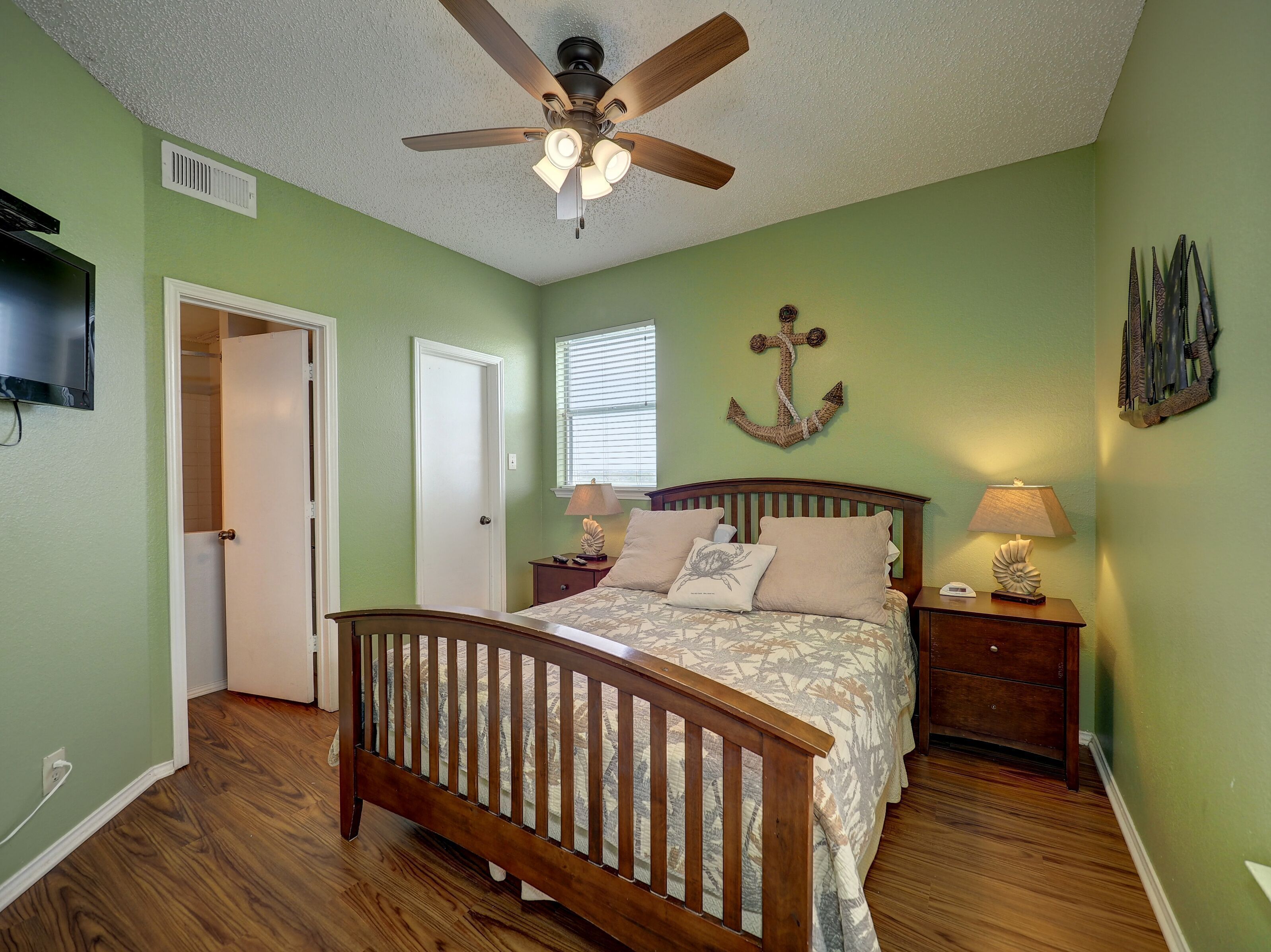 A bedroom with a wooden bed, a ceiling fan, a nightstand with a lamp, and a wall-mounted anchor decoration.