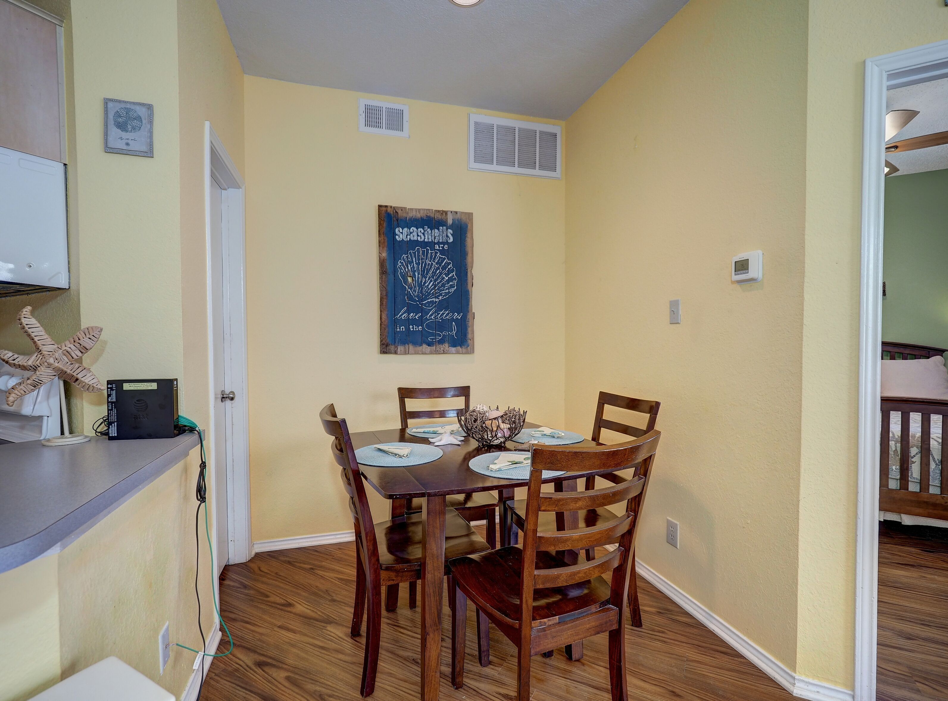 A dining area with a wooden table and chairs, a kitchen area with a white refrigerator, and a wall with a framed sign.
