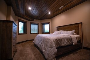 Ein Schlafzimmer mit Holzbalkendecke, einem großen Bett mit gemusterten Textilien und drei Fenstern mit Jalousien.
