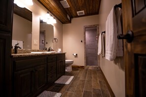 Ein Badezimmer mit Holzbalkendecke, einem großen Spiegel, einem Waschbecken mit Granitplatte, einer Toilette und einem Handtuchhalter.