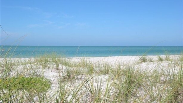 Una spiaggia con sabbia e poca erba, un cielo azzurro limpido e un mare calmo.