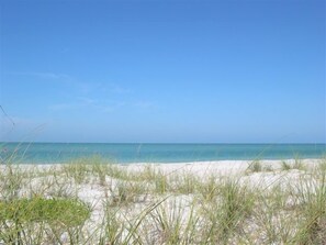 A beach with sand and sparse grass, a clear blue sky, and a calm sea. - Beautiful Palm Island House! Walk2Beach. Pool, Spa, Kayaks, Bikes, Dock.Amazing! (Palm Island)
