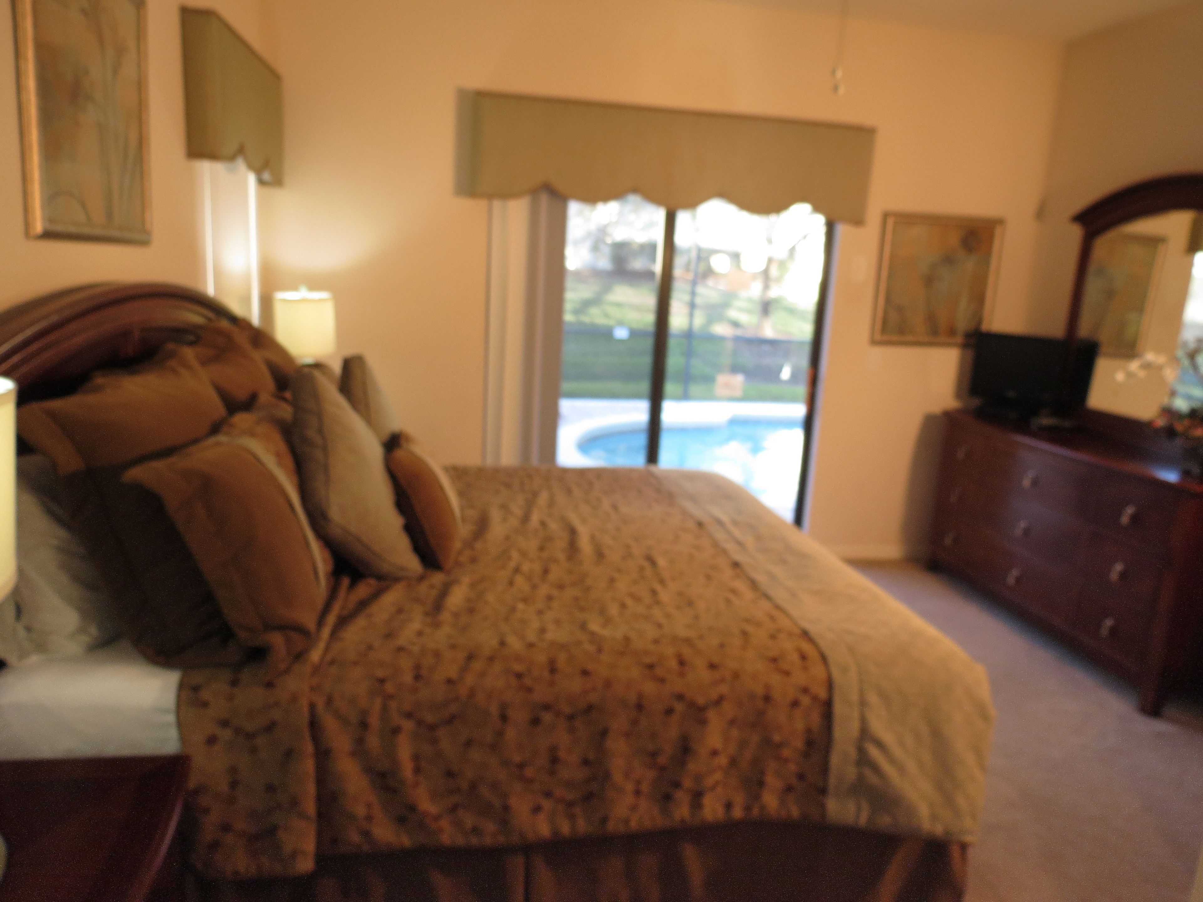A bedroom with a bed, a dresser, a TV, and a window with a view of a swimming pool.