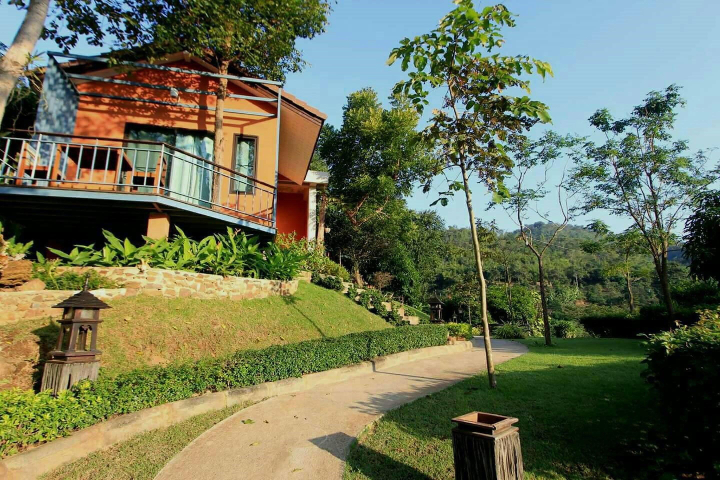 A modern house with a balcony, surrounded by lush greenery and a stone wall.