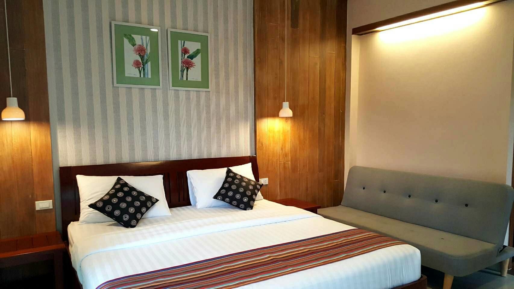 A hotel room with a bed, a grey sofa, two wall-mounted lamps, and two framed floral pictures.