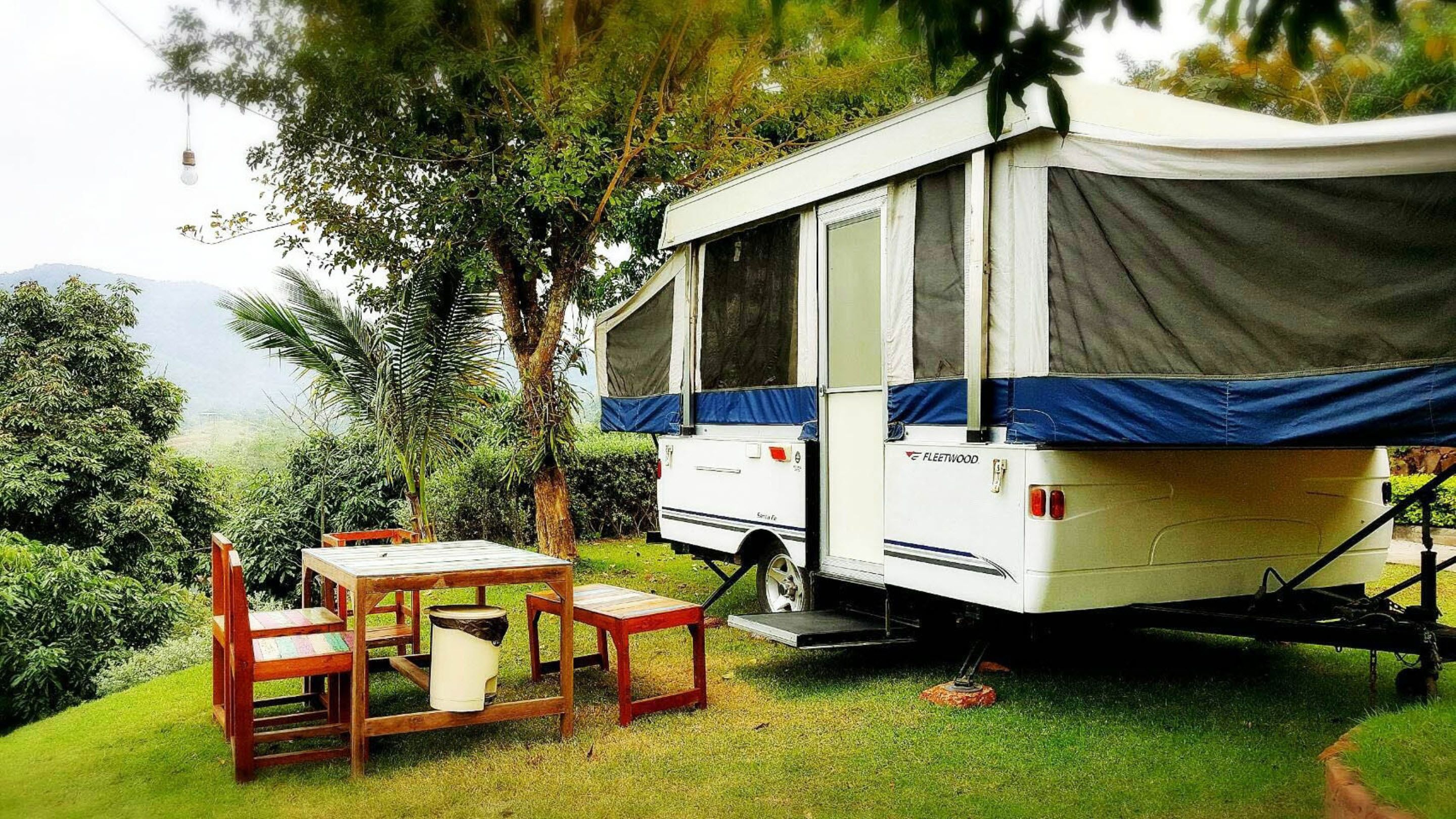 Una caravana blanca con detalles azules, una mesa, sillas y un pequeño contenedor de basura en un área verde con árboles y montañas al fondo.
