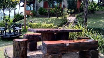 An outdoor seating area with rustic wooden benches and tables, surrounded by lush greenery and a building in the background.