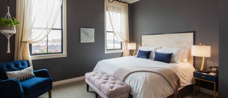 A bedroom with a large bed, two bedside tables, a blue armchair, and a hanging plant.