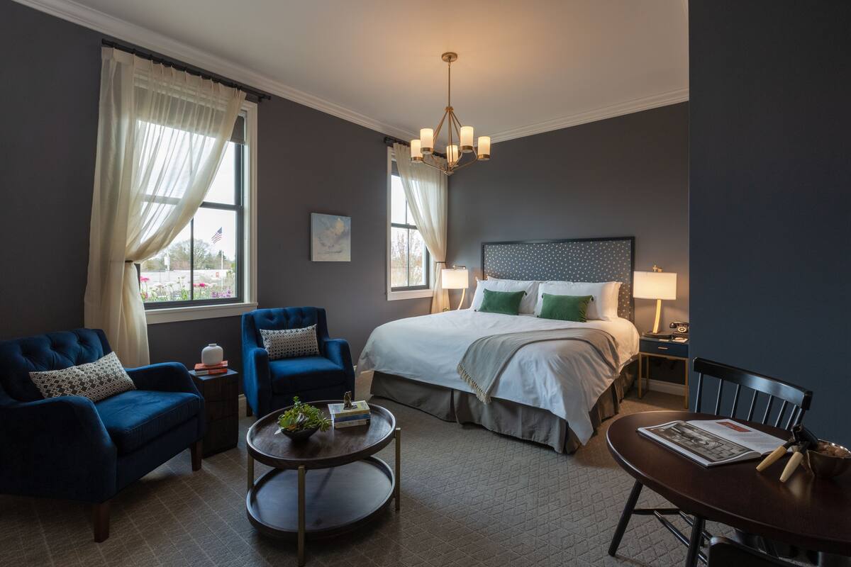 A hotel room with a large bed, two blue armchairs, a round coffee table, and a dining table with chairs.