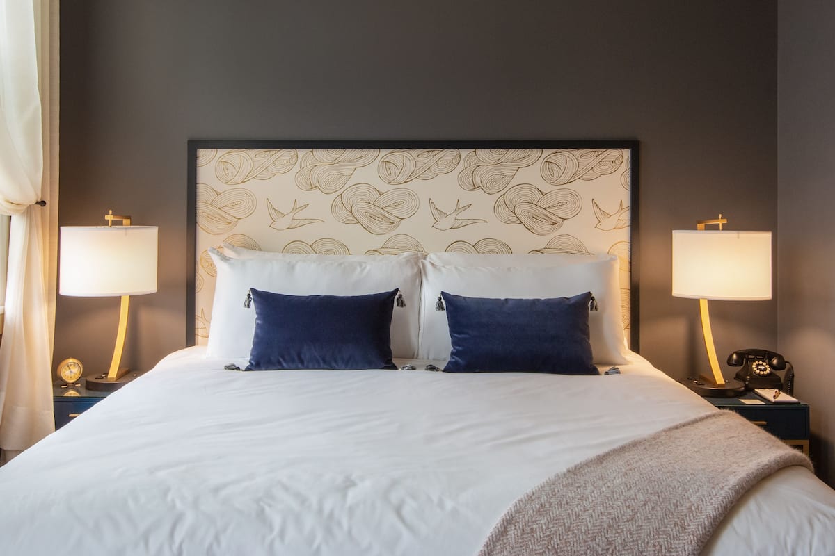 A neatly made bed with white linens and two blue pillows, flanked by nightstands with lamps and clocks.