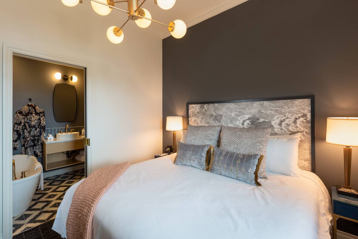 A bedroom with a large bed, a nightstand with a lamp, a dresser, a mirror, and a bathroom with a bathtub and sink.