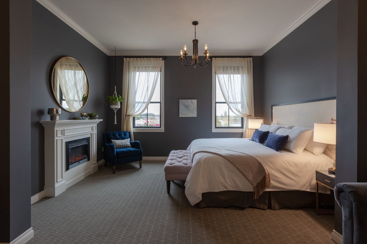A bedroom with a large bed, a fireplace, a blue armchair, and a chandelier.