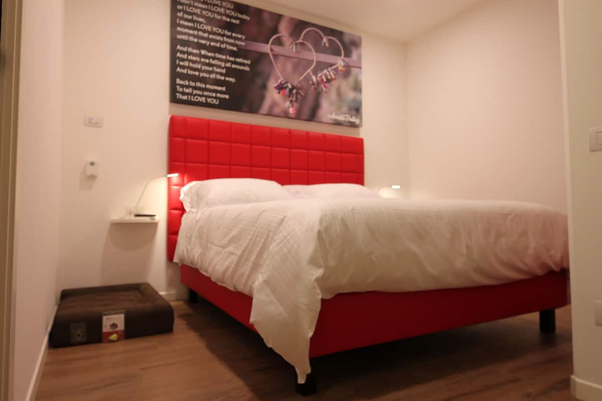 A bedroom with a red headboard, a white bedspread, a bedside table with a lamp, and a poster on the wall.