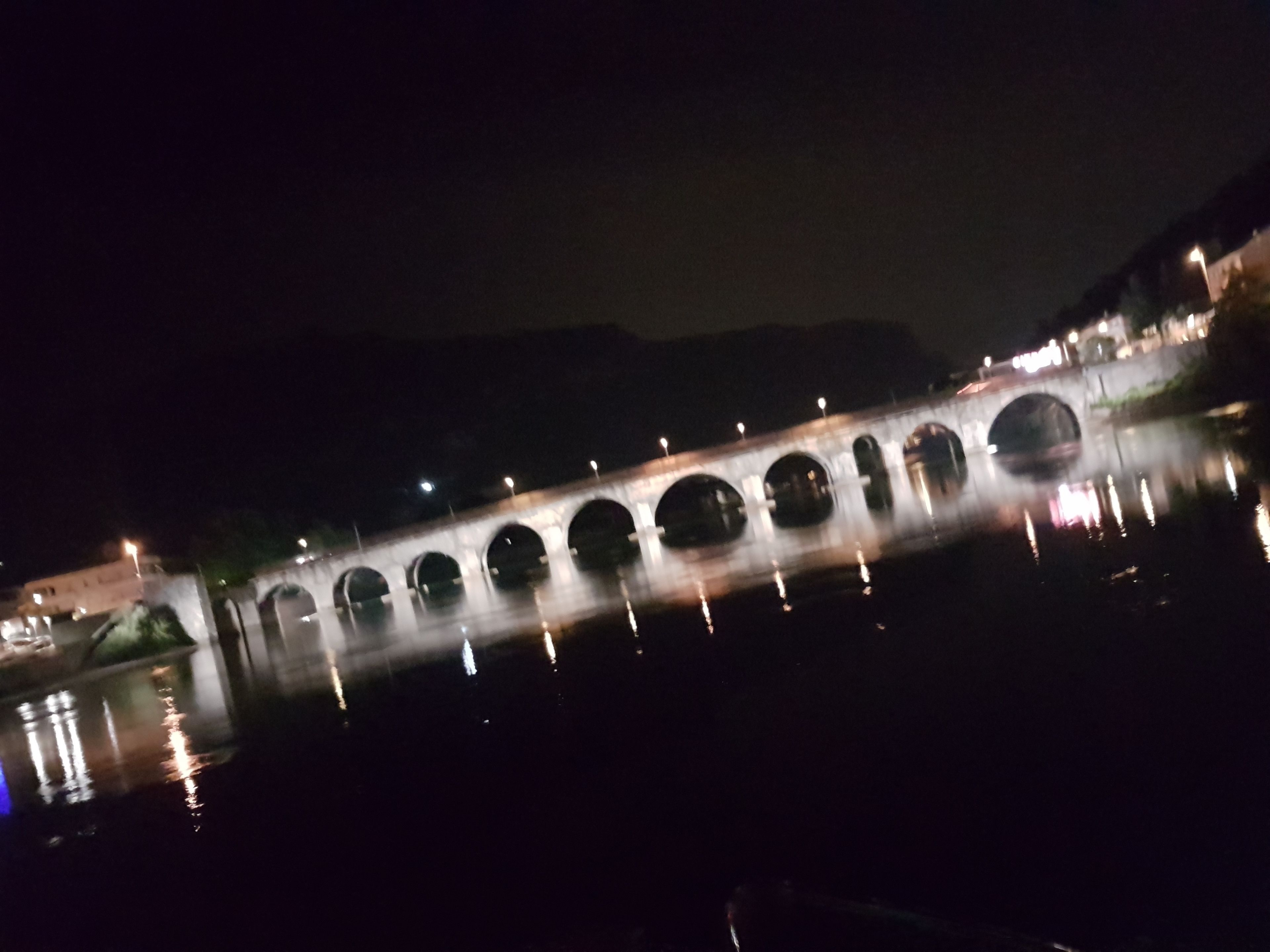 Une vue nocturne d’un pont à arches multiples enjambant une étendue d’eau, illuminé par des lampadaires.