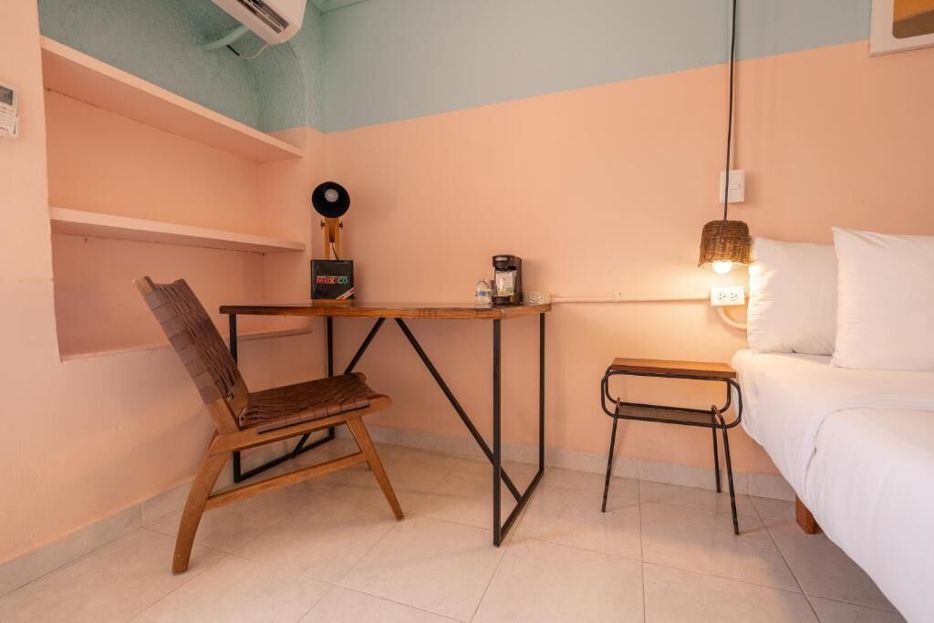 Una habitación pequeña y acogedora con una cama, una silla de madera, una mesita con cafetera y una lámpara.