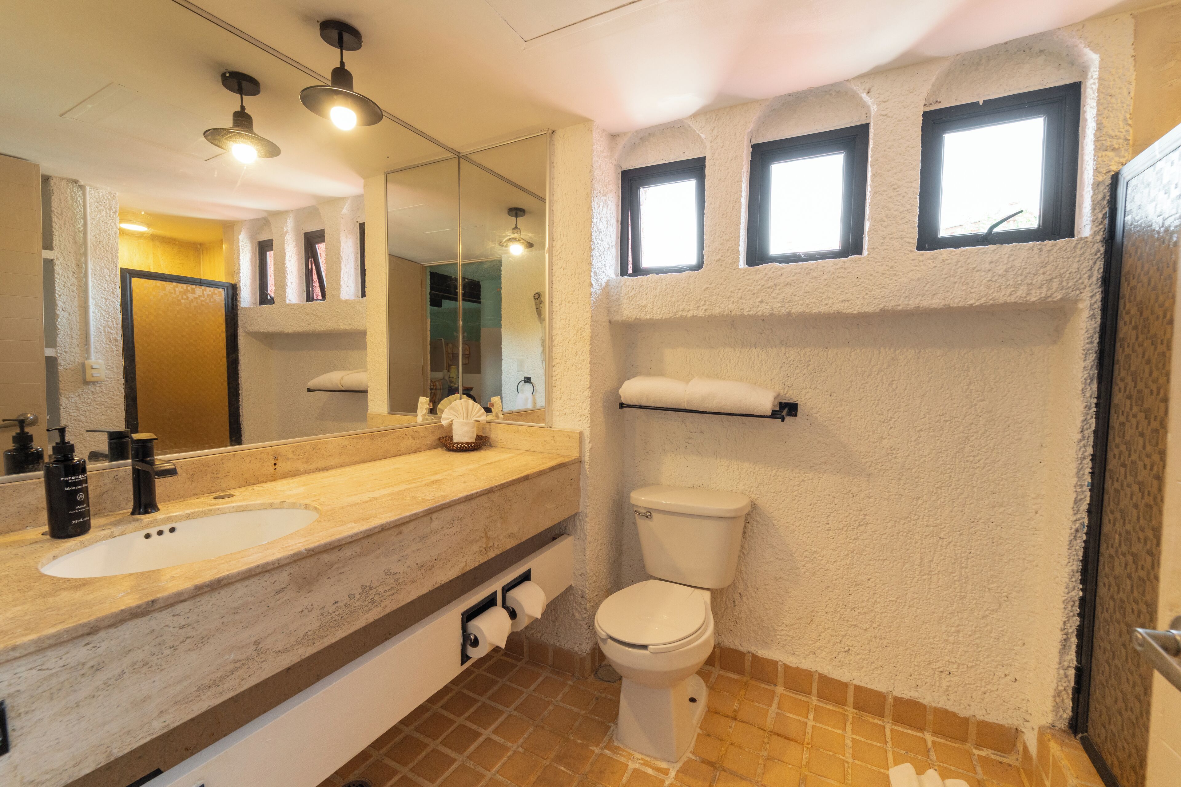 A bathroom with a marble sink, a toilet, and a mirror. There are three lights above the sink and two windows above the toilet.