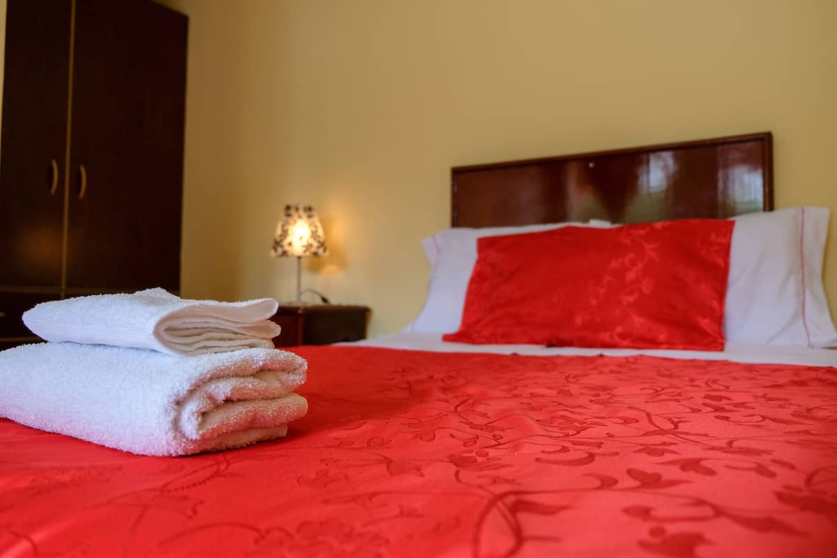 Una cama bien tendida con una colcha y una almohada rojas, una pila de toallas blancas y una mesita de noche con una lámpara.