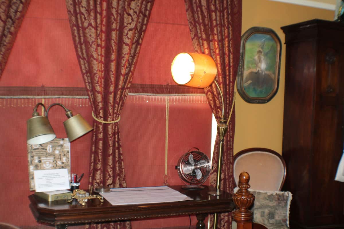 A desk with a lamp, a fan, and a chair in a room with patterned curtains and a wooden cabinet.