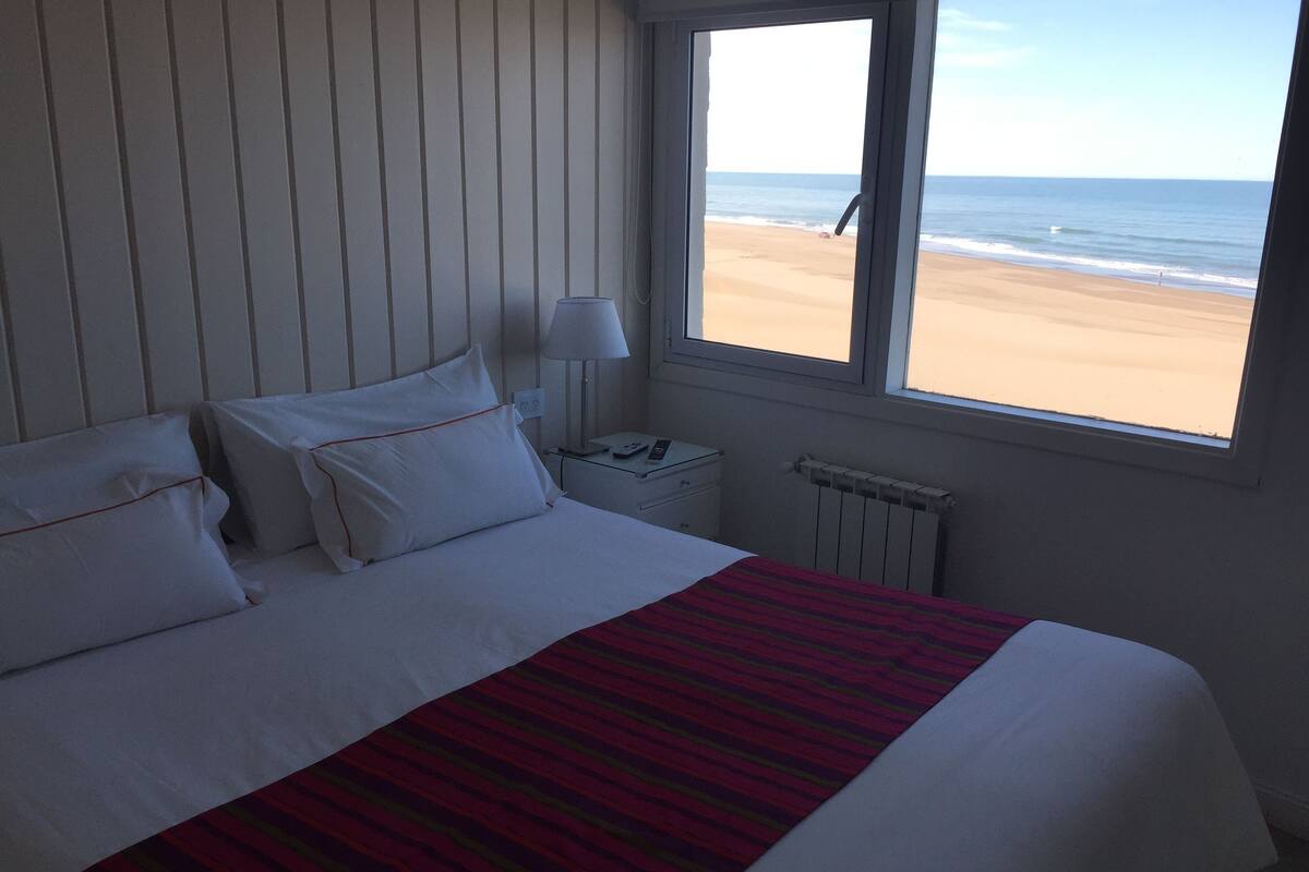 A hotel room with a bed, a nightstand, a lamp, and a window with a beach view.