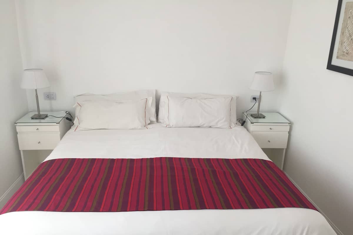 A neatly made bed with white bedding and a red and brown striped blanket. There are two bedside tables with lamps on either side of the bed.