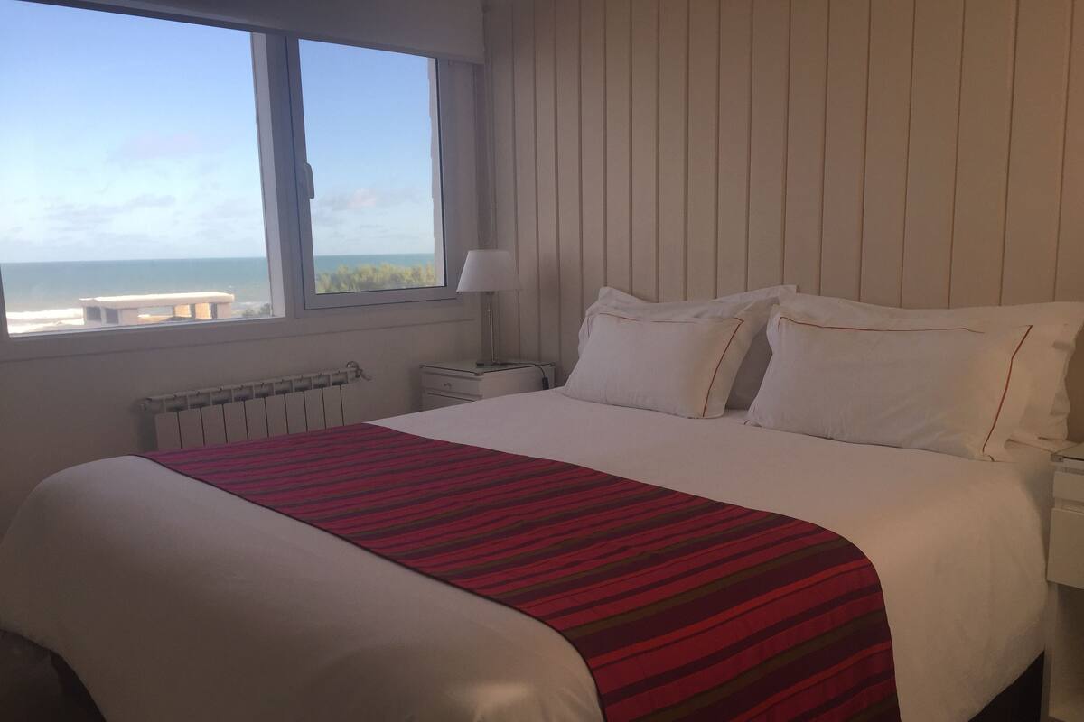 A hotel room with a large bed, a window with a view of the sea, a bedside table with a lamp, and a radiator.