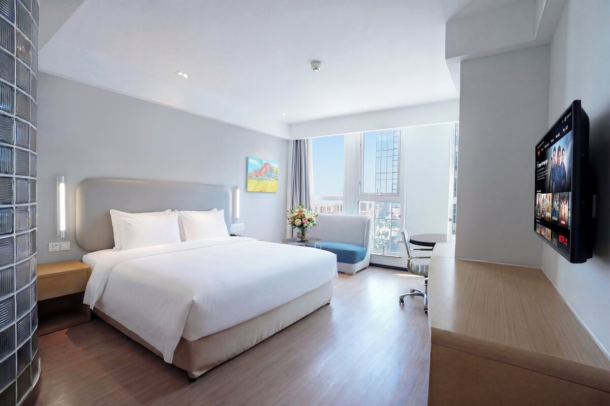 A modern hotel room with a large bed, a desk, a chair, a TV, and a city view.